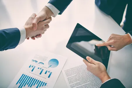 Symbolbild Vertrieb: Zwei Hände schütteln sich über einem Tisch mit einem Diagramm darauf, derweil wird ein Tablet bedient.