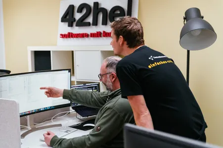 Axel und Arne schauen auf einen Bildschirm. Im Hintergrund das 42he-Logo.