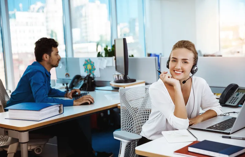 Support-Mitarbeiterin sitzt am Schreibtisch und lächelt in die Kamera. Im Hintergrund ein konzentrierter Kollege.