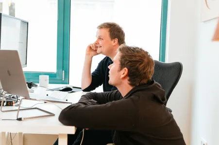 Christian und Axel schauen gut gelaunt auf einen Bildschirm, fotografiert von der Seite.