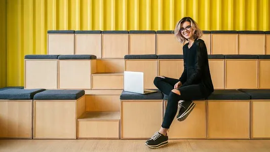 Eine Frau sitzt auf den Bänken einer Aula und lächelt in die Kamera. Neben ihr auf der Bank steht ein Laptop.