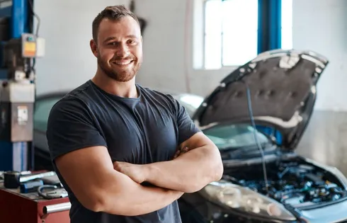 Handwerker posiert mit verschränkten Armen vor Motorhaube.